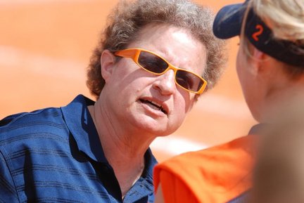 Coach Karla Wolters wearing orange sunglasses and a navy shirt, speaking to a softball player during a game. The background shows the softball field in warm sunlight.